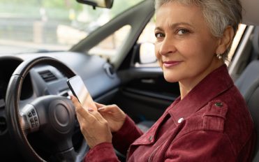 Portrait of elegant modern female pensioner using electronic gadget inside automobile. Stylish mature woman sitting in driver seat of vehicle she rented via car sharing application on her phone